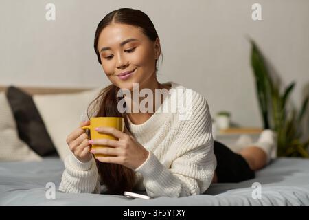 Una donna asiatica rilassata si gode una bevanda calda mentre si rilassa nel suo appartamento moderno Foto Stock