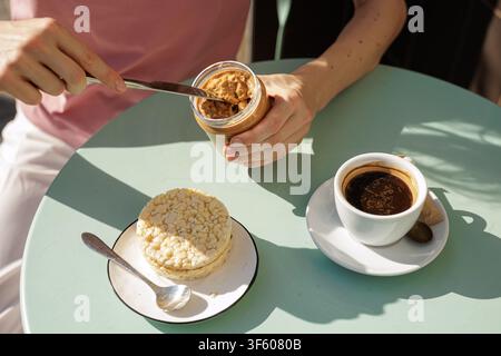 Una persona prepara torte di riso con burro di arachidi croccante mentre gusta una tazza di caffè caldo a un tavolo all'aperto, illustrando una colazione semplice ed equilibrata Foto Stock