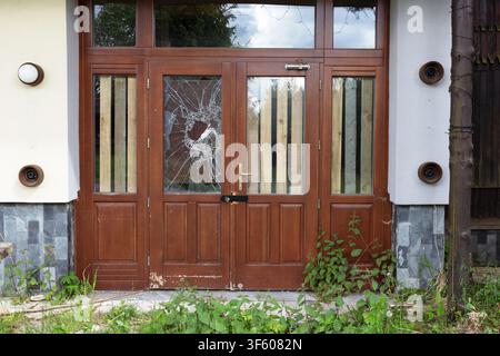 Ingresso abbandonato di un vecchio edificio trascurato con finestra rotta, vetri frantumati e vegetazione invasa. Architettura fatiscente, luogo dimenticato, u Foto Stock