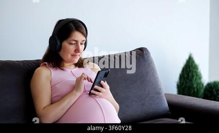 Donna incinta che si rilassa sul divano ascoltando musica con lo smartphone Foto Stock