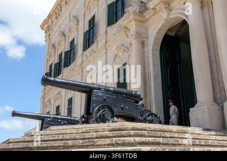 Cannoni storici affiancano l'ingresso barocco dell'Auberge de Castille a Valletta, Malta, una guardia in servizio alla porta dell'ufficio del Primo Ministro Foto Stock