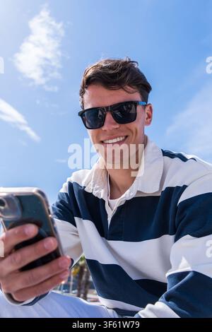 Giovane uomo che controlla il suo smartphone vicino a una marina, sorridendo nello sfondo soleggiato Foto Stock