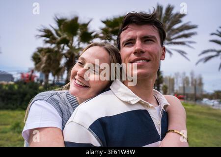 Coppia che si abbraccia calorosamente e sorride insieme in un parco, i capelli che volano nel vento in una giornata nuvolosa Foto Stock