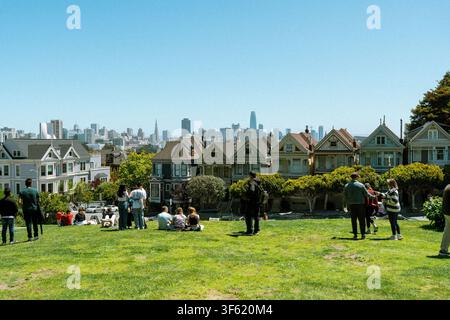 I visitatori si rilassano sul prato di Alamo Square Park, San Francisco, con le famose case vittoriane Painted Ladies e lo skyline del centro città alle loro spalle Foto Stock