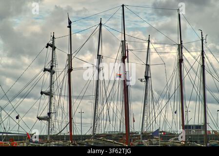 Alti alberi di navi e intricate sartie che creano un motivo nautico contro un cielo drammatico, con una bandiera arcobaleno colorata Foto Stock