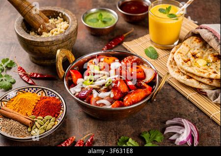 Pollo tikka indiano servito in una ciotola di metallo tradizionale con pane naan, lassi al mango, chutney alla menta, spezie e verdure fresche su un tavolo rustico. Foto Stock