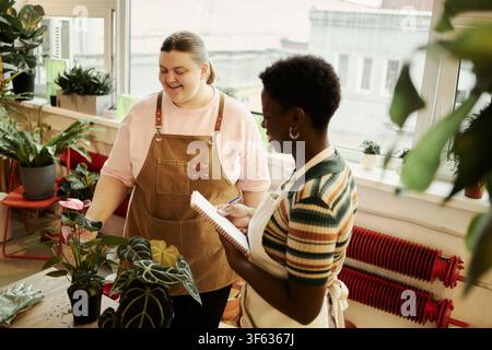 Giovani fioriste diverse che lavorano in un negozio di piante, una donna caucasica sorride mentre una collega afroamericana prende appunti. Scatto ad angolo alto che rappresenta il lavoro di squadra nelle piccole imprese Foto Stock