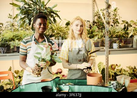 Ritratto di una giovane donna afroamericana e di una giovane donna caucasica che tengono piante in vaso in una serra. L'immagine rappresenta la proprietà di una piccola impresa e il marketing di uno stile di vita ecologico Foto Stock