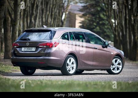 Berlino, Germania - 8 aprile 2026: Opel Astra K Sports Tourer station wagon 2016 5a generazione 1.6 BiTurbo auto marrone parcheggiata su un vialetto di ghiaia, percorso di pista, vicolo, alberi, retro Foto Stock