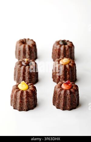 Primo piano di sei canelés assortiti (tortine al rum) con ripieni di cioccolato, caramello salato, crema al limone e crema al lampone su sfondo bianco Foto Stock