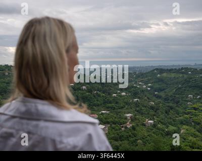 Donna bionda in piedi di spalle alla macchina fotografica, che guarda le colline verdi lussureggianti coperte di case sparse, con il cielo nuvoloso, le città distanti Foto Stock