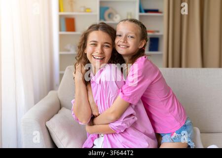 Madre e figlia allegre che si abbracciano strettamente sul divano e sorridono alla telecamera Foto Stock