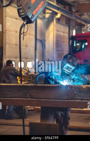 Vista verticale di un saldatore professionista con maschera protettiva che salda una trave d'acciaio con scintille blu in un'officina dell'industria pesante Foto Stock