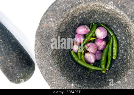 Mortaio e pestello tradizionali in pietra di granito con scalogni freschi e peperoncini verdi rossi, vista dall'alto su sfondo bianco, pronti per preparare una pasta asiatica piccante. Foto Stock