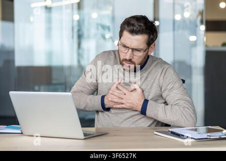 Giovane uomo d'affari che avverte dolori toracici acuti o sintomi di infarto, si stringe il petto con entrambe le mani mentre è seduto alla scrivania, sopraffatto dallo stress lavorativo Foto Stock