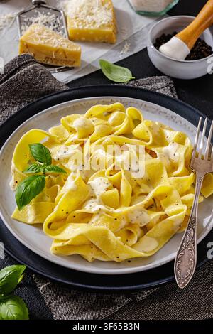 Pasta Tagliatelle Alfredo ricoperta di salsa al formaggio cremosa, pepe nero e basilico fresco su piatto con forchetta. Pezzi di parmigiano, grattugia e mortaio Foto Stock
