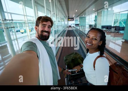 Coppia felice che scatta un selfie viaggiando sul marciapiede mobile del terminal dell'aeroporto. Foto Stock