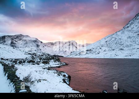 Trascinante tramonto invernale che getta un bagliore rosa sulle cime innevate delle montagne che circondano il lago Llyn Ogwen a Eryri (Snowdonia), nel Galles del Nord. Foto Stock