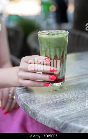 Mano che tiene un bicchiere alto di latte al matcha verde con una manicure rosa brillante su un tavolo esterno ruvido accanto a sedute morbide da caffè e piante, con matcha Foto Stock