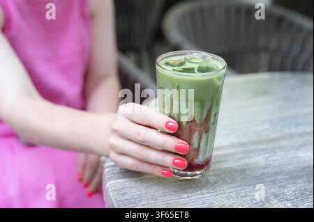 Giovane donna che tiene un latte matcha ghiacciato accanto a un tavolo da caffè all'aperto con una manicure vivace rossa e rosa, bicchiere pieno di latte vorticoso, ghiaccio e crema Foto Stock