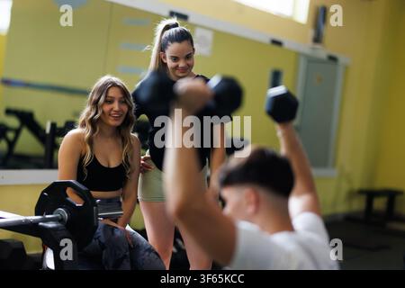 Due donne osservano un uomo che solleva pesi in una palestra durante il giorno mentre si impegnano in attività di fitness e sostengono la sua routine di allenamento Foto Stock