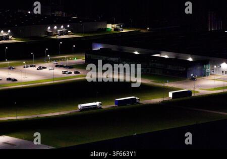 Vista aerea di camion che percorrono una strada vicino a un magazzino industriale e un parcheggio luminosamente illuminati di notte a Vilnius, contea di Vilnius, Lituania. Foto Stock