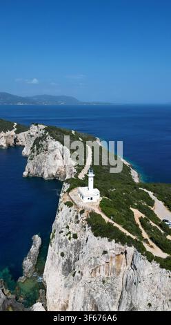Stupenda ripresa verticale con drone di un faro bianco storico in cima a un'alta scogliera rocciosa. Mare blu profondo bellissimo, strada sterrata costiera e cespugli verdi Foto Stock