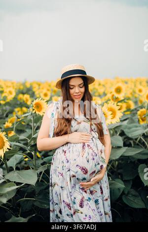 Giovane donna incinta che tocca delicatamente la pancia con un sorriso pacifico. Una futura mamma che posa all'aperto in un campo in fiore, concetto di gravidanza e maternità. Foto Stock