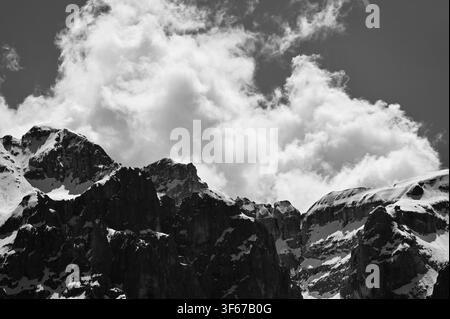 Una fotografia di paesaggio drammatica in bianco e nero che cattura picchi montuosi frastagliati, ricoperti di neve, che si innalzano bruscamente sotto enormi nuvole cumulo che si gonfiano. Il trattamento monocromatico ad alto contrasto enfatizza le trame ruvide dei pendii rocciosi e le forme morbide e organiche del cielo drammatico. Foto Stock