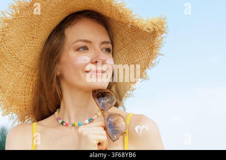 Donna sorridente con cappello di paglia, crema solare sul viso e sulla spalla, tiene occhiali da sole, promuovendo la protezione SPF, la cura idratante della pelle e la prevenzione dal sole Foto Stock