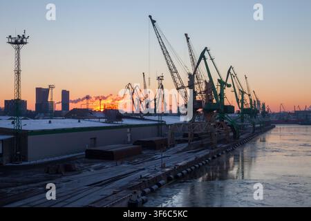Porto industriale all'alba con gru per il carico lungo un lungomare ghiacciato, cielo invernale, binari ferroviari e magazzini che creano una scena di porto di lavoro freddo. Foto Stock