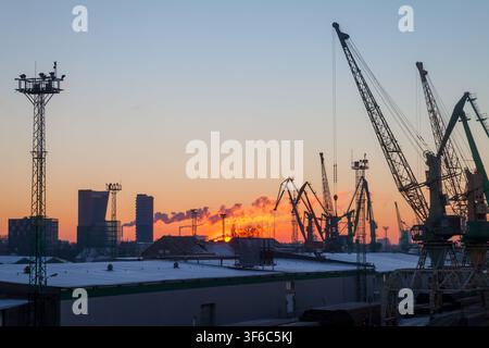 Porto industriale all'alba con gru per il carico lungo un lungomare ghiacciato, cielo invernale, binari ferroviari e magazzini che creano una scena di porto di lavoro freddo. Foto Stock