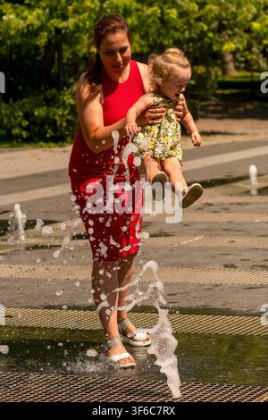 La madre solleva rapidamente la sua figlia piccola spaventata sopra getti d'acqua schizzanti mentre la porta attraverso una fontana da terra all'aperto in una giornata soleggiata. Foto Stock