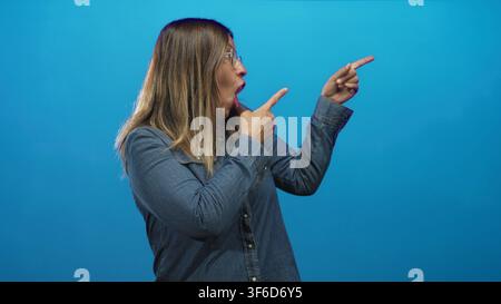Donna di mezza età indica con entrambe le dita verso destra con bocca aperta e occhiali, indossa una camicia di jeans in uno studio blu; reazione di sorpresa. Foto Stock