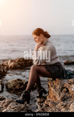 Donna oceano tramonto Giovane donna seduta pensierosa su scogli sul mare durante l'ora dorata contemplando la vita Foto Stock