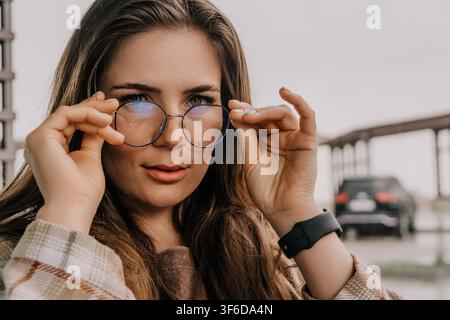 Ritratto di donna con occhiali giovane donna che regola eleganti occhiali rotondi all'aperto contro uno sfondo sfocato morbido della città Foto Stock