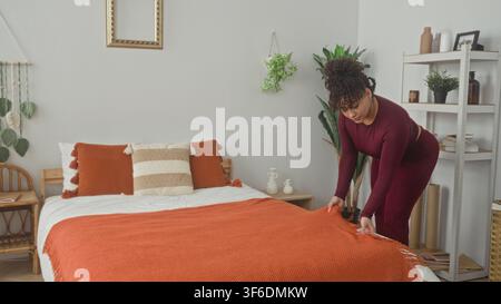 Donna che nasconde una coperta arancione sul letto con le mani e i cuscini liscianti mentre si piega vicino allo scaffale in una camera da letto dell'edificio; calma routine. Foto Stock