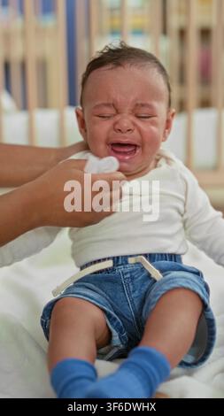 Bambino ispanico che piange, tiene un sonaglio di legno mentre mani adulte asciugano le lacrime in una culla di edificio; cure di consolazione in caso di distress. Foto Stock