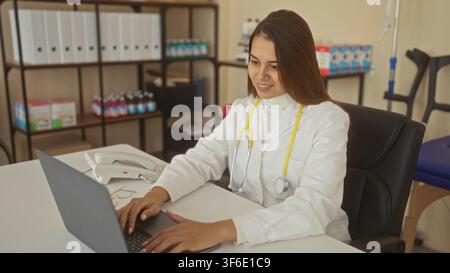 Dottoressa che digita su un laptop alla scrivania in un edificio, stetoscopio al collo con occhiali e telefono sul tavolo; cure concentrate. Foto Stock
