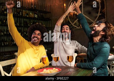 Gruppo multirazziale di amici maschi che guardano una partita di calcio al bar. Celebrare un punteggio, con pugni in su Foto Stock