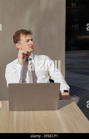 Giovane uomo d'affari in camicia bianca e cravatta seduto a un tavolo all'aperto con un laptop, guarda pensieroso di lato mentre lavora alla luce calda del sole vicino a un mo... Foto Stock