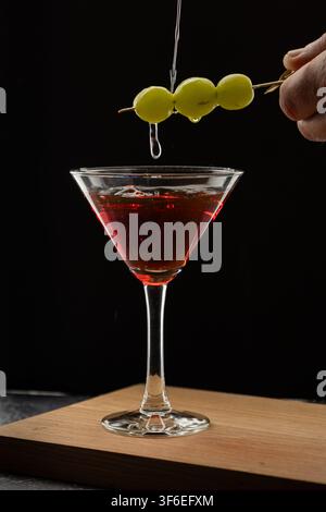 Una mano tiene uno spiedino con tre acini d'uva verde che gocciolano sciroppo su un bicchiere da martini pieno di cocktail rosso scuro, su uno sfondo nero su legno Foto Stock