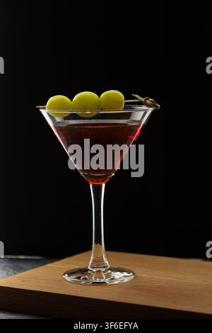 Tre acini d'uva verde su uno spiedino appoggiati sul bordo di un bicchiere da martini pieno di cocktail rosso scuro, fotografati su uno sfondo nero in legno Foto Stock
