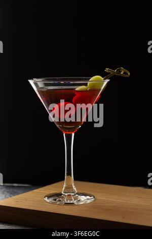 Un bicchiere da martini classico riempito con un cocktail rosso intenso si trova su una tavola di legno contro uno sfondo nero, con una luce rossa drammatica che si riflette lungo la Foto Stock