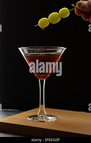 Una mano sospende uno spiedino con tre acini d'uva verde con una singola goccia che si forma sopra un bicchiere da martini con cocktail rosso scuro su una tavola di legno. Foto Stock