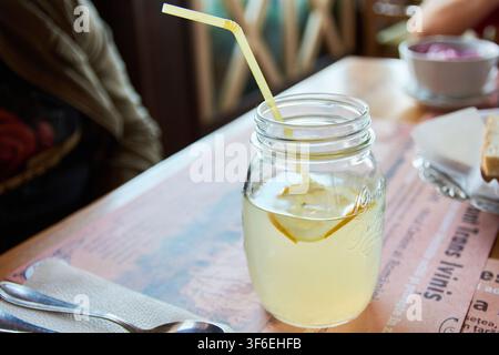 Primo piano di una limonata fredda e rinfrescante in un barattolo di vetro trasparente con una cannuccia gialla, servita su un tavolo di ristorante con altri piatti e utensili. Foto Stock
