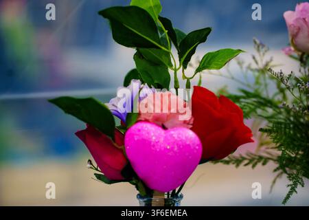 Un mazzo di fiori tra cui rose rosse, garofani rosa e tocchi viola è disposto in un vaso. Un cuore rosa decora il bouquet, rendendolo speciale. Foto Stock