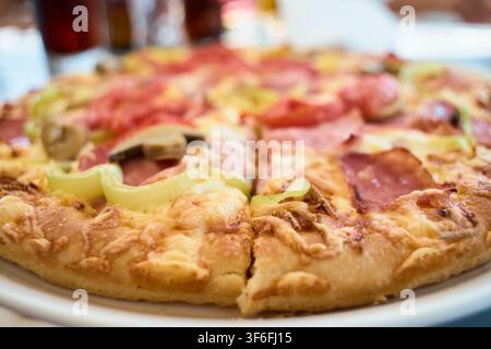 Primo piano di una pizza fatta in casa appena sfornata con formaggio fuso, funghi affettati, prosciutto e peperoni, servita su un piatto bianco. Foto Stock