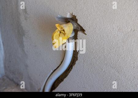 Un tubo di plastica bianco con un raccordo metallico è parzialmente visibile e fuoriesce da un buco frastagliato in un muro ruvido, fissato con nastro adesivo giallo Foto Stock