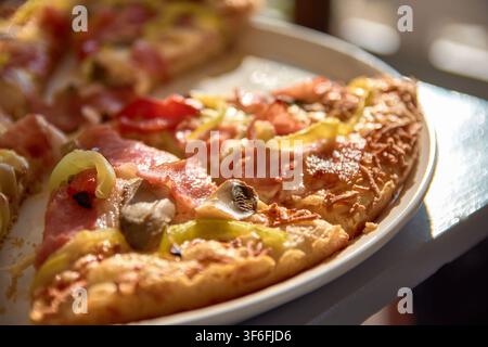 Una vista ravvicinata di fette di pizza appena sfornate ricoperte di formaggio fuso, prosciutto, funghi e peperoni, servite su un piatto bianco. Foto Stock